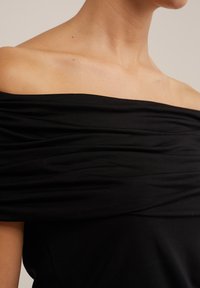 Maglia nera scollata sulle spalle con tessuto arricciato, texture liscia e design aderente che valorizza il décolleté e le spalle.