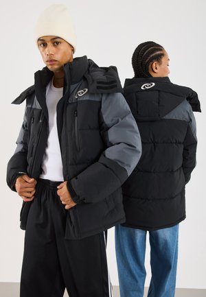 Veste rembourrée en noir et gris, dotée d'une fermeture éclair à l'avant, de poches latérales et d'un col montant. Capuche ajustable avec un détail de marque.