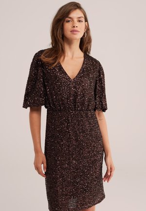 Femme portant une robe brun foncé à sequins avec manches courtes et col en V, debout devant un fond clair uni.