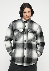 Camicia in flanella a quadri bianchi e neri con due tasche sul petto, chiusura con bottoni e una consistenza morbida. Orlo arrotondato e maniche lunghe.