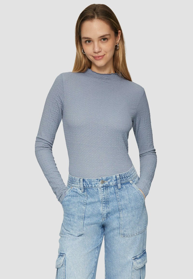 Hellblaues Langarmoberteil aus strukturiertem Stoff mit hohem Kragen, kombiniert mit hochgeschnittenen hellblauen Cargo-Hosen aus Denim mit Taschen.