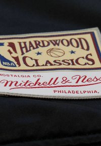 Etichetta in tessuto blu navy con la scritta "HARDWOOD CLASSICS" in crema e rosso, un'icona di basket e "Mitchell & Ness" in corsivo, e "PHILADELPHIA."