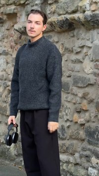 Tamno siva pletena sweater s visokim ovratnikom, u kombinaciji s labavim crnim hlačama. Osoba drži crne slušalice, stojeći uz kameni zid.