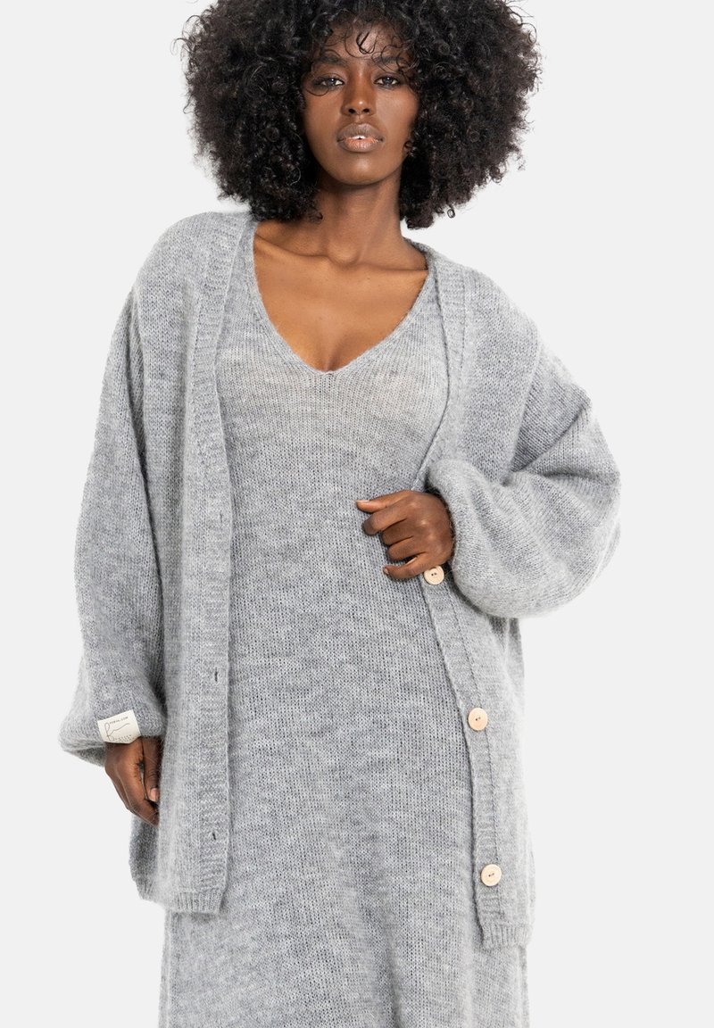 Szary sweter na drutach z dużymi rękawami, z drewnianymi guzikami i teksturowanym wykończeniem, noszony na szarą sukienkę o dopasowanym kolorze.