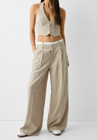 Chaleco corto beige con botones y escote halter combinado con pantalones beige de piernas anchas con una cintura blanca y ligera textura.