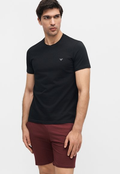 Άνδρας φορώντας μαύρο κοντομάνικο μπλουζάκι με μικρό λογότυπο στο στήθος και μαρόν σορτς, στέκεται σε λευκό φόντο.