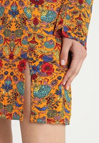 Vestido de estampado floral colorido en naranja, con mangas cortas, una abertura lateral y patrones intrincados en tonos rojos, azules y verdes.