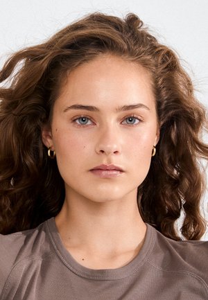 Jeune femme aux cheveux bruns bouclés, yeux bleus, portant de petites boucles d'oreilles en or et un t-shirt marron à col rond, regardant droit devant avec une expression neutre.