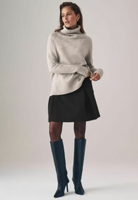 Hellgrauer Rollkragenpullover mit geripptem Saum, kombiniert mit einem schwarzen Faltenrock und kniehohen schwarzen Lederstiefeln. Einfaches und strukturiertes Design.