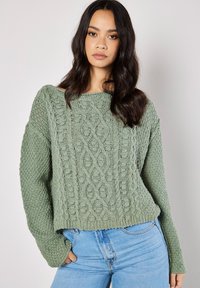 Flauschiger Strickpullover in gedämpftem Grün mit strukturierten Mustern, weit geschnittenem Bootsausschnitt und langen, locker sitzenden Ärmeln, kombiniert mit hellblauen Jeans.