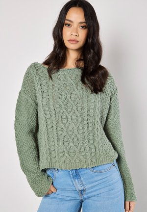Chunky gebreide trui in gedempt groen met textuurpatronen, een brede boothals en losse lange mouwen, gecombineerd met lichtblauwe jeans.