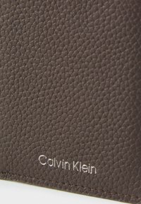 Ruskea teksturoitu nahkalompakko, jossa on kohokuvioitu "Calvin Klein" -logo hopeisena. Sen pinnassa on sileä, hieman kiiltävä tekstuuri ja hienovaraiset saumat.