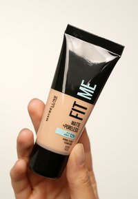 Podkład Maybelline Fit Me w przezroczystej tubce, w kolorze brzoskwiniowym, z czarną zakrętką. Posiada wyraźny napis oraz małą niebieską etykietę wskazującą na 12-godzinne trwałość.