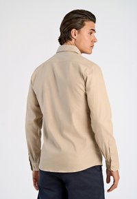 Camicia beige a maniche lunghe con colletto classico, dettaglio della spalla sul retro e polsini con bottoni. Realizzata in tessuto morbido con una texture liscia.