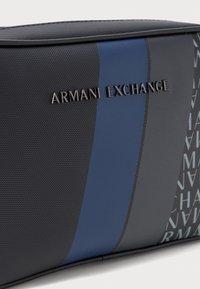 Juodas ir mėlynas tekstūruotas Armani Exchange krepšys su vertikaliomis juostelėmis ir prekės ženklo logotipu metaliniais ženklais ant priekinio skydelio.