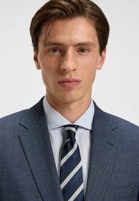 Heren suit jacket van een gestructureerde, donkerblauwe stof, gecombineerd met een lichtblauw gestreept shirt en een donkerblauw met witte streepjes stropdas.