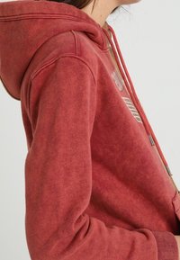 Hoodie in cotone rosso con tasca a marsupio frontale, cordino regolabile e design stampato sul petto. Tessuto morbido, vestibilità comoda.