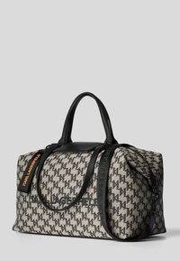 Sac de sport beige et noir avec motif pied-de-poule, accents en cuir, deux poignées supérieures et une sangle amovible avec étiquette de logo orange.