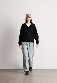 Μαύρη φούτερ ζακέτα με κολάρο που έχει φερμουάρ, ανοιχτό γαλάζιο φαρδύ τζιν και παπούτσια με σχέδια. Το μοντέλο φοράει μπεζ καπέλο και λευκό μπλουζάκι από κάτω.