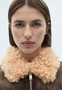 Jachetă din suede maro, cu un guler curly pufoș, portocalie deschis și o fermoar argintie, pe un fundal albastru deschis.