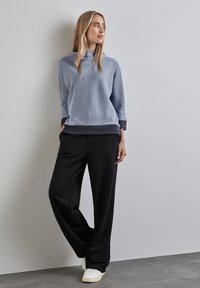 Felpa azzurra chiara con motivo e vestibilità comoda, abbinata a pantaloni neri a gamba larga e scarpe da ginnastica bianche, in piedi contro uno sfondo neutro.