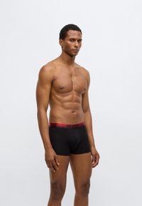 Boxer noir avec une ceinture élastique rouge portant le nom de la marque "HUGO" en lettres noires. Tissu lisse et coupe ajustée.