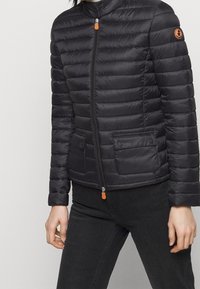 Sort pufferjakke med horisontalt quiltet design, lynlås foran, høj krave og to flaplommer med knap. Har orange lynlåsdrag og logo.