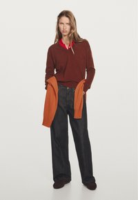 Maglione marrone su una camicia rossa con colletto, abbinato a jeans di denim scuri. Un cardigan arancione è appoggiato sul braccio. La texture sembra morbida.