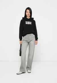 Hoodie preto com texto branco "TOMMY JEANS", combinado com jeans claros de perna larga e sapatos beges. Modelo leva uma pequena mala preta.