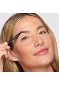 Femme aux cheveux châtain clair utilisant un pinceau biseauté pour appliquer du maquillage sur son sourcil droit, montrant un gros plan de son visage.