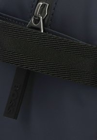 Bolsa de nailon negra con correa texturada negra, que presenta un cierre de cremallera plateado y una etiqueta de marca negra. Superficie lisa con acabado mate.