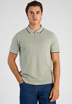 Polo met korte mouwen in lichtgroen, voorzien van een ritssluiting aan de voorkant, een kraag met marineblauwe en witte bies, en een gladde textuur. Draagt bij blauwe spijkerbroek.