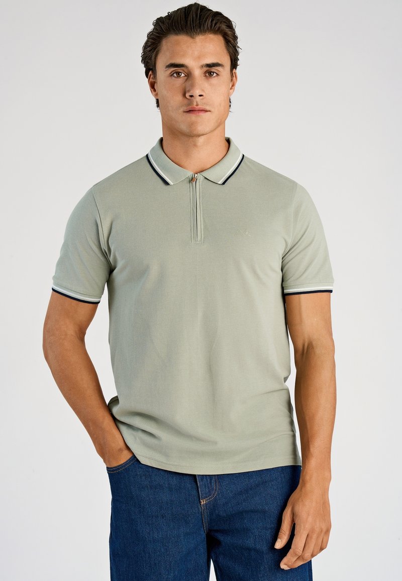 Koszulka polo z krótkim rękawem w jasnym zielonym kolorze, z zamkiem błyskawicznym z przodu, kołnierzykiem z granatowo-białym wykończeniem oraz gładką fakturą. Nosi się ją z niebieskimi jeansami.