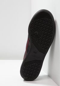 Suela negra de una zapatilla Adidas mirando hacia adelante, mostrando el patrón de textura de la suela y el logotipo de la marca, apoyada contra una superficie blanca de esquina.