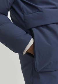 Marinblå puffjacka med bälte, har en strukturerad yta och en kontrasterande vit manschett. Dold sidofickdetalj synlig.