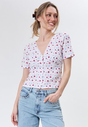 Smilende kvinde med blondt hår i en hestehale ifører sig en hvid blomsterknappet bluse og lyseblå jeans med bølget lommedetalje.