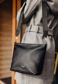 Černá kožená crossbody taška s hladkým povrchem, obdélníkovým tvarem a stříbrnými detaily. Obsahuje logo a pevný popruh.