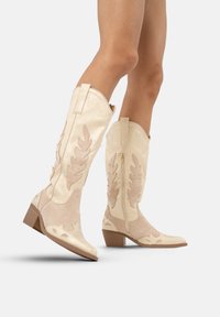Botas cowboy hasta la rodilla en ante color crema y beige, con recortes en forma de hoja, un zipper lateral y un tacón apilado para mayor estabilidad.