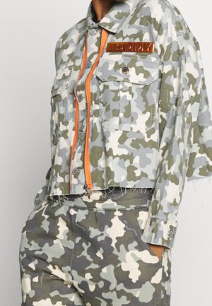 Veste camouflage courte avec fermeture éclair orange et poche poitrine, associée à un pantalon camouflage assorti avec poches latérales, main visible dans la poche.
