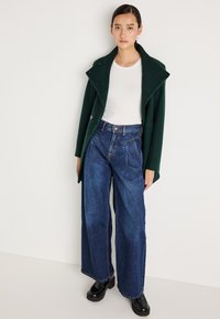 Casaco verde em mistura de lã com um colarinho largo e fechamento com botão duplo; combinado com jeans de ganga escuros de perna larga e cintura alta, e botas de plataforma pretas.