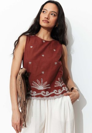 Femme portant un haut rouille sans manches avec broderie blanche et pantalon blanc, tenant une pochette beige à franges, debout sur fond blanc.