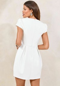 Robe blanche courte avec un col rond, des manches courtes et des plis au niveau de la taille. Elle possède une fermeture éclair discrète au dos et une texture de tissu lisse.