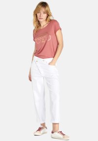 Roze T-shirt met gouden bedrukte tekst, samen met een losse witte broek en lichtgekleurde sneakers met rode accenten.