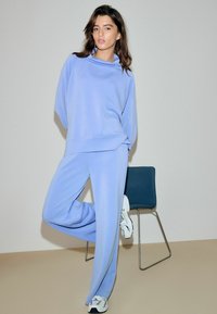 Lyseblåt athleisure sæt bestående af en sweatshirt med løs pasform og matchende vidde bukser, lavet af blødt, glat stof. Hvide sneakers og en minimalistisk stol fuldender looket.