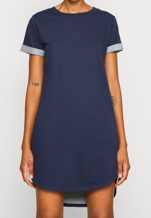 Femme portant une robe bleu marine à manches courtes avec des poignets gris retroussés et des tatouages visibles sur les deux avant-bras et bras supérieurs.