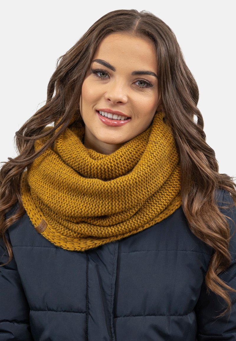 Bufanda infinita de punto en color mostaza, con un patrón texturizado, llevada sobre una chaqueta azul marino. Presenta una pequeña etiqueta de marca marrón.