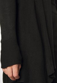 Černý pletený kardigan s žebrovanou texturou, dlouhými rukávy a splývavým střihem. Materiál je měkký, lehký a poskytuje pohodlný volný střih.