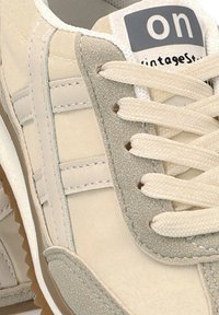Zapatilla beige claro con texturas de ante y cuero. Presenta cordones blancos, acentos grises y un logo "vintageStyle" en la lengüeta.