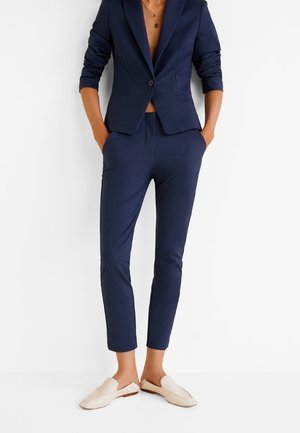 Femme portant un blazer bleu marine cintré et un pantalon slim assorti avec des chaussures beige à enfiler, mains dans les poches, sur fond blanc uni.