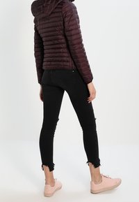 Chaqueta acolchada color burdeos con capucha, vaqueros skinny negros desgastados y zapatillas rosas, vista desde atrás, mostrando una silueta ajustada.